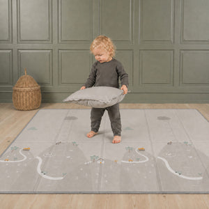 A toddler standing and playing safely in a living room on the shock-absorbing grey landscape side of the Allis play mat.