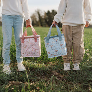 Two children walking outdoors carrying Little Dutch cooler bags in pink Fairy Garden and blue Forest Friends designs.