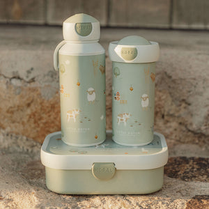 The Little Farm school beaker displayed outdoors on a stone surface, sitting next to a matching water bottle on top of a matching light green lunchbox.