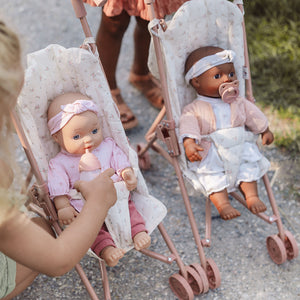 Close-up of two baby dolls sitting comfortably strapped into Little Dutch doll strollers outdoors.