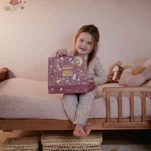 A young girl with light brown hair sits on a wooden bed holding a decorative Little Dutch pink story projector flashlight box. She is wearing a light pink and white patterned pajama set.