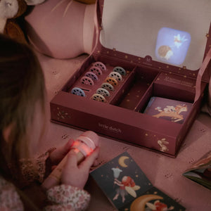 A child holds a small glowing fairy light in a cozy bedroom setting, with an open 'little dutch story projector flashlight gift box showing a set of colorful discs and booklet on a pink bedspread.