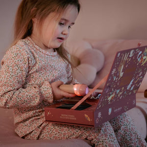 A young child in floral pajamas sits on a couch holding a flashlight over an open little dutch story projector box. The scene is softly lit and cozy, with the child as the main subject in a real indoor environment.