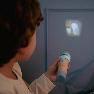 A child holds little dutch story projector flashlight with a light blue and beige body, shining a projected image of a white bird onto a gray wall in a dimly lit room.