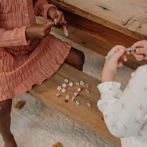 Two children sitting at a rustic wooden table, holding and examining small pastel gemstone beads scattered on the bench. One child wears a dusty rose ruffled dress, and the other wears a white floral long-sleeve top.