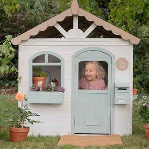 Children's playhouse with a child peeking out of the door, surrounded by plants and flowers.