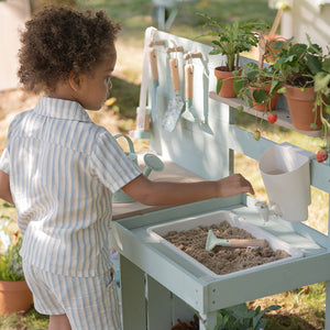 A young boy using a toy shovel to mix sand and mud inside the sink basin of a Little Dutch outdoor mud kitchen.