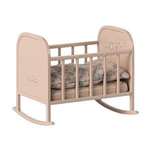 A miniature metal cradle in light rose color with a floral pattern on the headboard, gently curved legs, and bedding inside.