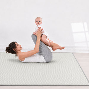 A woman in a white tank top and gray leggings lies on a light gray textured yoga mat, holding a baby who is sitting on her knee in a bright, minimalist room.