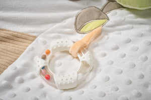 Baby teething ring on a textured white blanket with a soft toy in the background