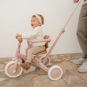 Toddler securely seated in the Little Dutch pink push tricycle with the safety ring attached, guided by a parent outdoors.