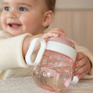 A smiling baby easily gripping the lightweight Little Dutch pink Fairy Garden spout cup by its ergonomic white side handles.