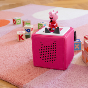 Lifestyle image showing the Peppa Pig Tonie figure placed on top of a pink Toniebox, sitting on a striped peach and pink mat with scattered wooden alphabet blocks in the background.