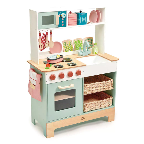 A large tender leaf toys kitchen with an oven, microwave, sink, hob, drawers, and shelves. The kitchen also includes a pot holder, plates, utensils, a fish dish with garnishes, herbs, and a tea towel.