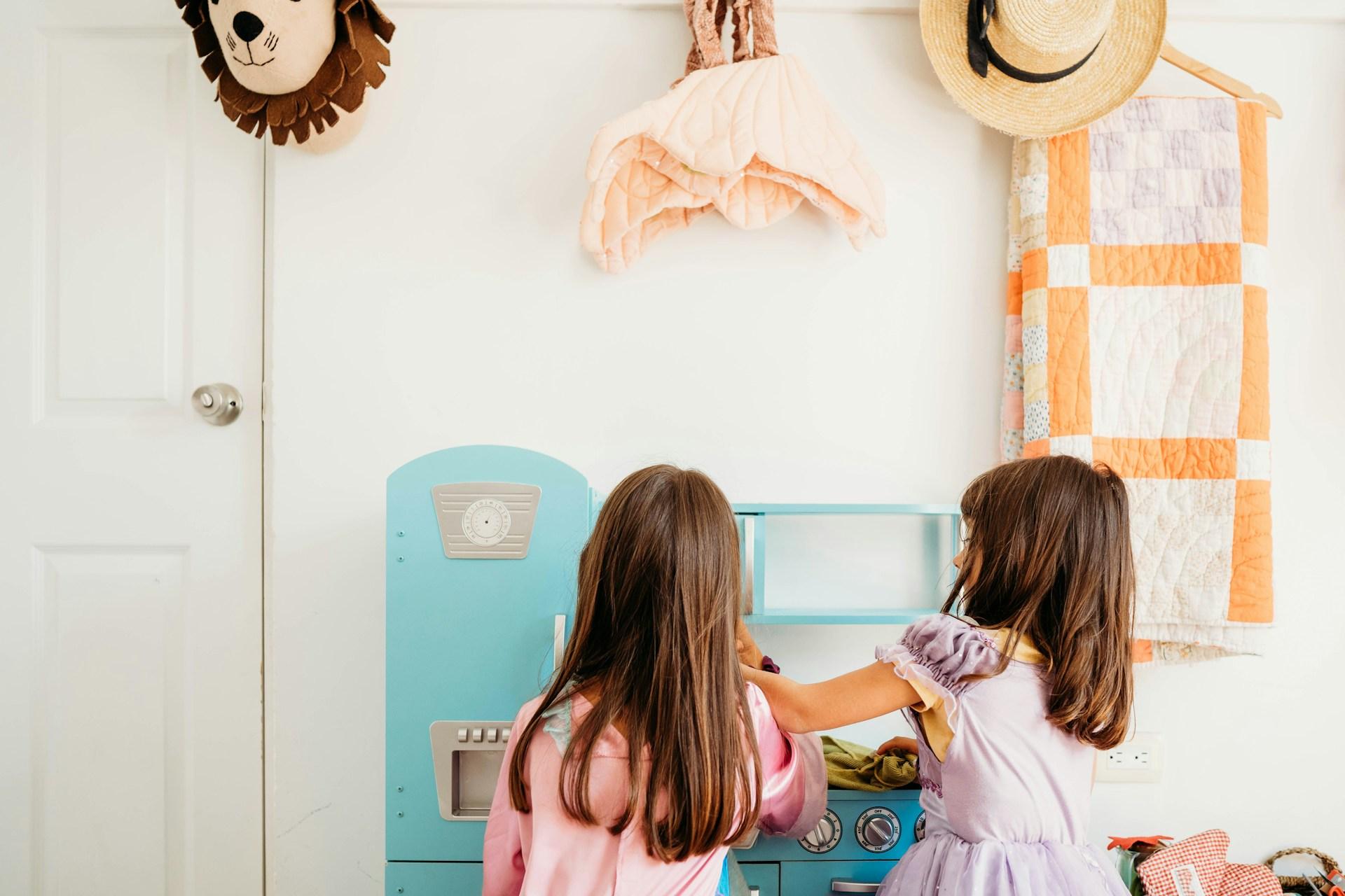 Two children playing together at a blue wooden play kitchen with fridge and oven, demonstrating cooperative role play