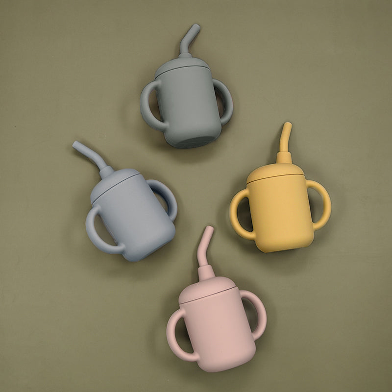 Overhead view of four silicone sippy cups in grey, mustard, pink, and olive green on a sage green background.