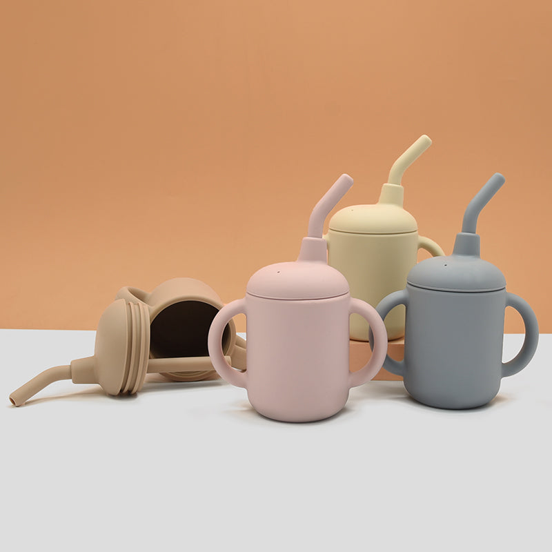 Three silicone toddler sippy cups in pink, blue, and yellow styled on a warm terracotta background.