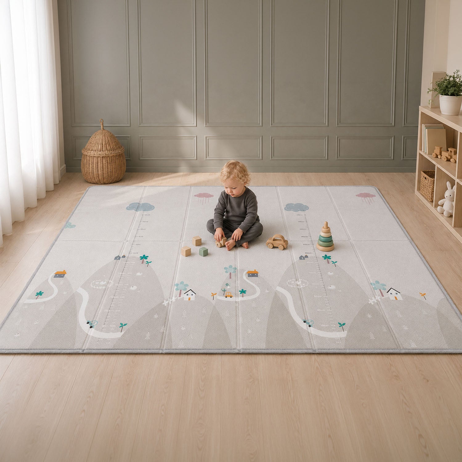 A toddler sits on a large fold-out play mat in a bright room with light wood floors and paneled walls. The play mat has a soft gray and white design with illustrated roads, clouds, trees, houses, and measurement markings.