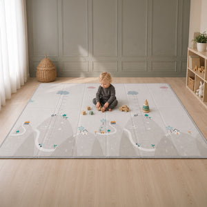 A toddler sits on a large fold-out play mat in a bright room with light wood floors and paneled walls. The play mat has a soft gray and white design with illustrated roads, clouds, trees, houses, and measurement markings.