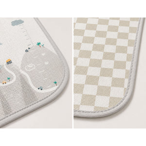 Two close-up views of a soft rectangular mat with rounded corners and stitched edges, shown side by side. The left view shows a light gray textured surface with a small illustrated road, palm tree, and vehicle motif.