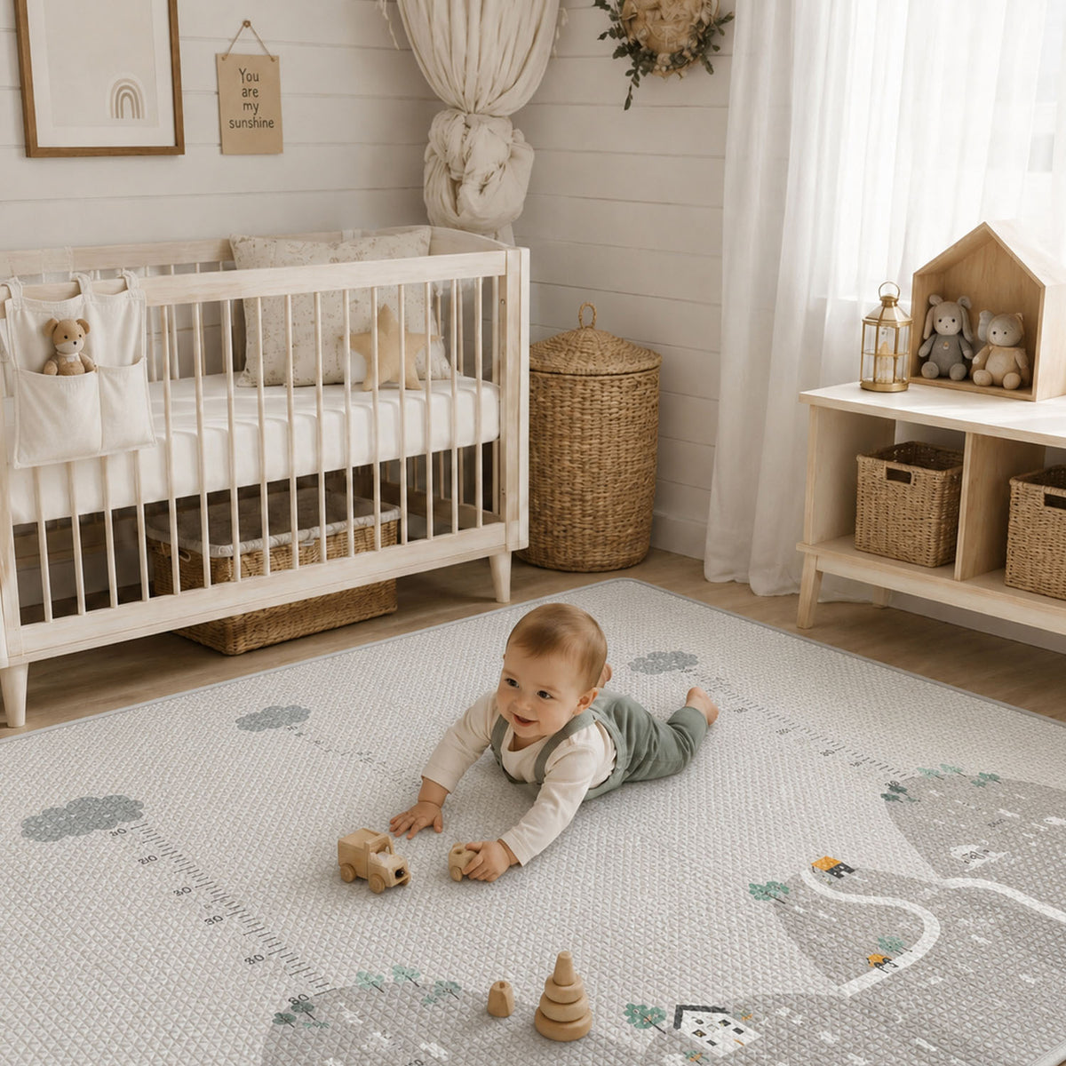 A baby lies on a light gray and white play mat with a road and cloud pattern in a styled nursery. The room includes a white crib, woven baskets, a wooden shelf with toys, and soft neutral decor.
