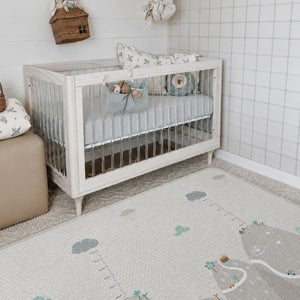 A white wooden baby crib with a light blue fabric hanging pouch on the front, styled in a bright nursery with a patterned rug, white tiled walls, and a small ottoman.