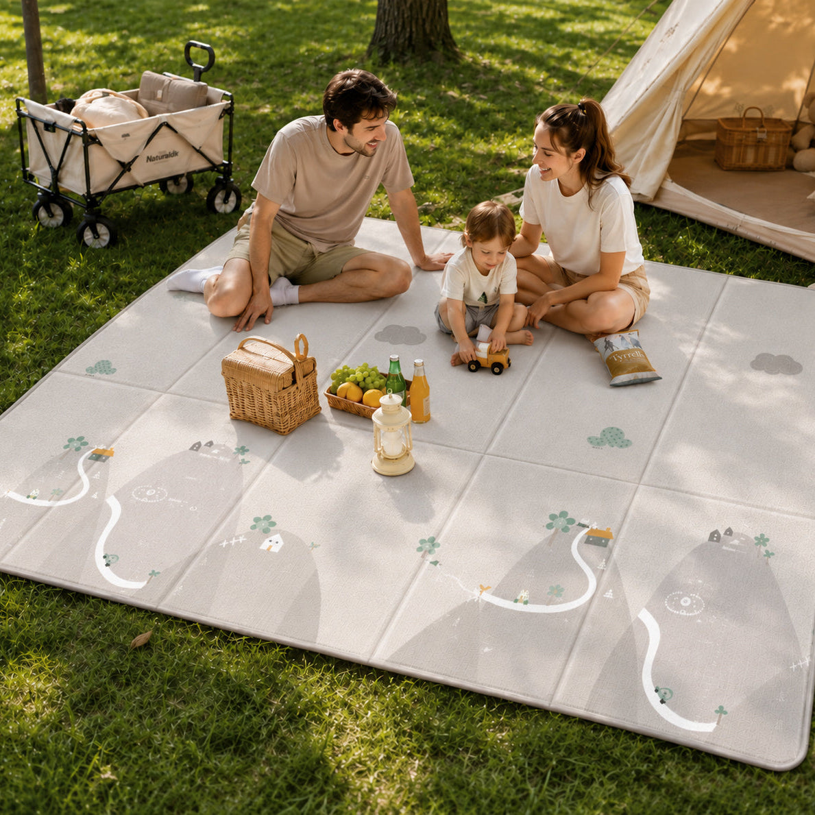 A family of three sits on a large gray outdoor play mat with cloud and animal illustrations on a grassy lawn. A man, a woman, and a young child are arranged on the mat with picnic accessories, toys, a wicker basket, and a small tent in the background.