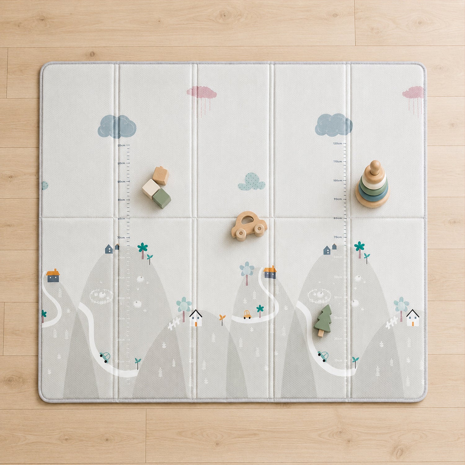 A large foldable children's play mat laid flat on a light wood floor. The mat has a soft gray and white mountain landscape print with clouds, small houses, trees, and measurement markings.