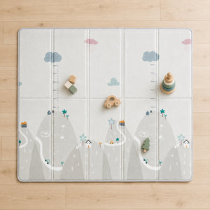 A large foldable children's play mat laid flat on a light wood floor. The mat has a soft gray and white mountain landscape print with clouds, small houses, trees, and measurement markings.