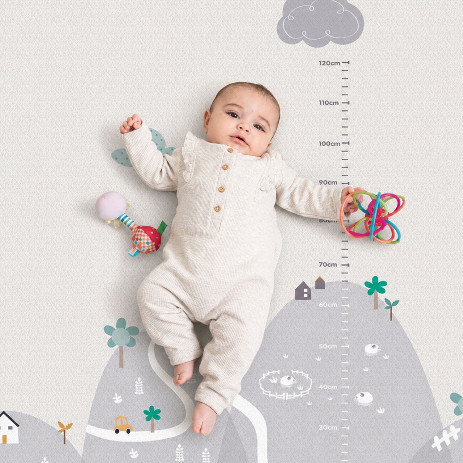A baby lies on a gray and off-white height measurement wall decal featuring a mountain, trees, houses, and a cloud. The wall sticker includes centimeter height markings from 30cm to 120cm.