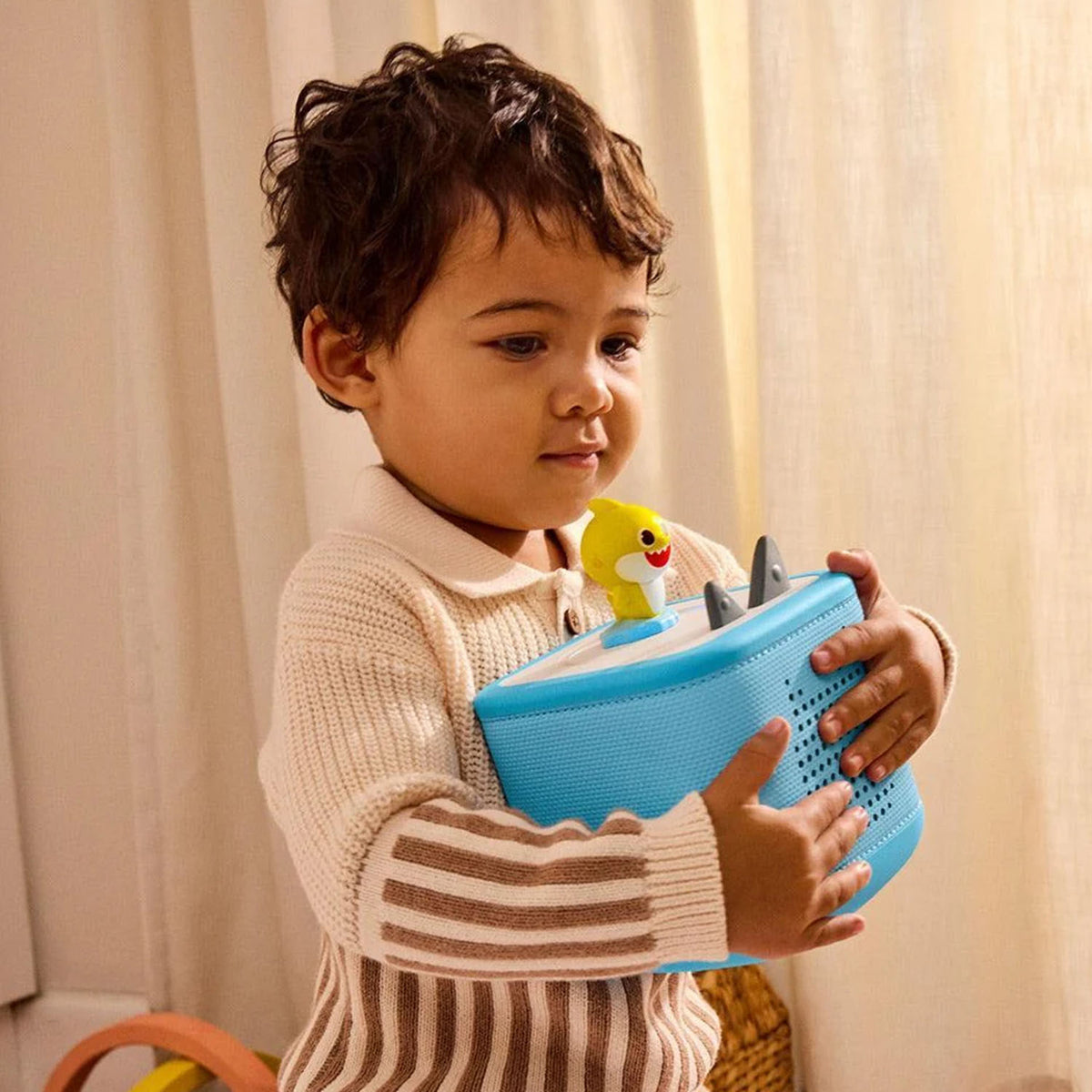 A young child smiling while holding a light blue Toniebox speaker, playing the Tonies Baby Shark audio figure.