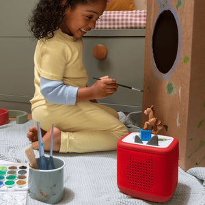 A young child playing in a cardboard playhouse in the background while the Tonies Toy Story 5 Bullseye audio figure sits on top of a red Toniebox speaker in the foreground.