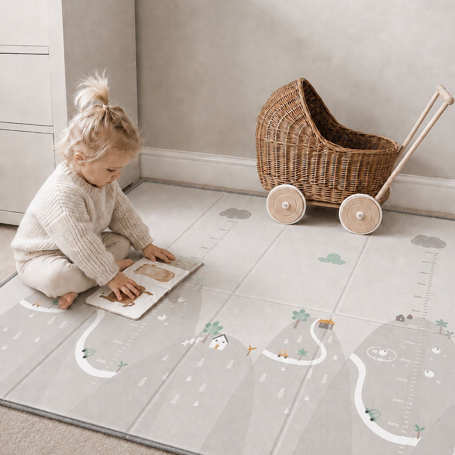 A toddler sits on a large gray floor mat with printed road, trees, and measurement markings, playing with a wooden book. A wicker baby carriage toy is pushed against the wall in the background.