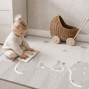 A toddler sits on a large gray floor mat with printed road, trees, and measurement markings, playing with a wooden book. A wicker baby carriage toy is pushed against the wall in the background.