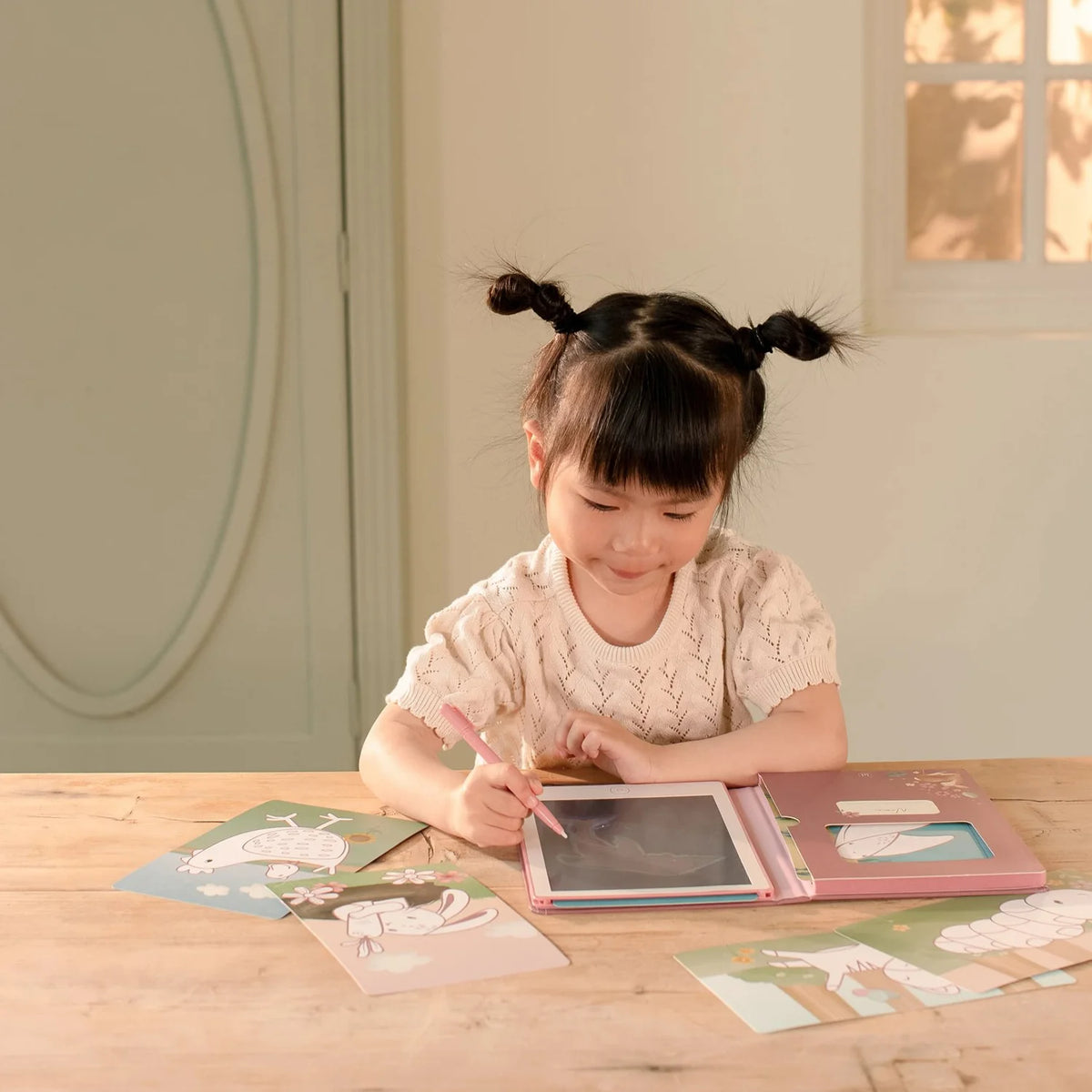 Young girl drawing on Little Dutch Pink Fairy Garden tablet at table with inspiration cards