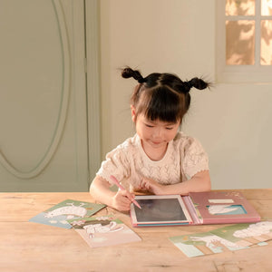 Young girl drawing on Little Dutch Pink Fairy Garden tablet at table with inspiration cards