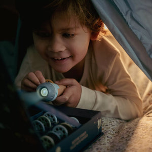 A young child smiling while holding a small Little Dutch flashlight under a tent, with a box of projectors.
