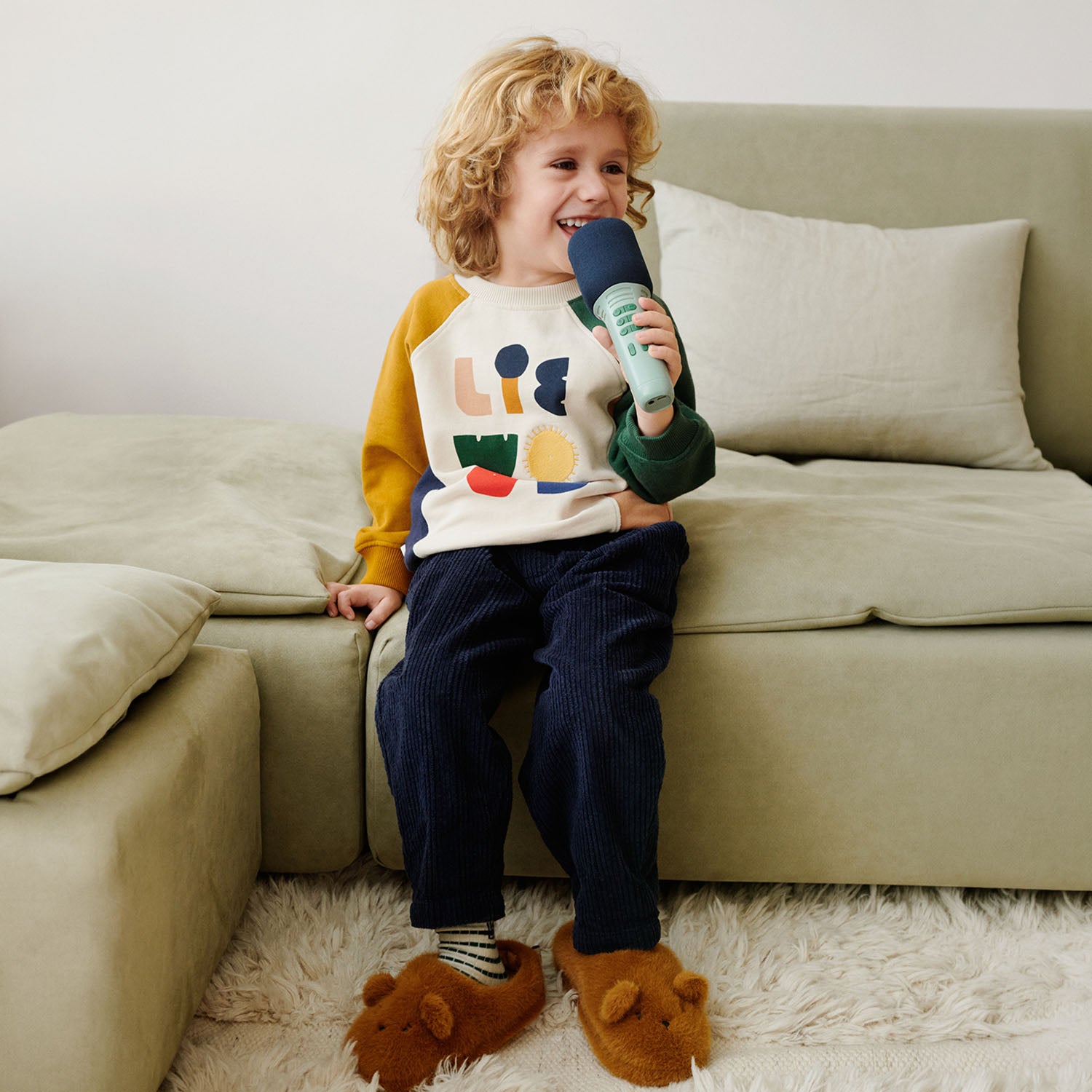 A young child sitting comfortably on a sofa, wearing a colorful sweater, dark trousers, and warm bear slippers.