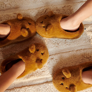 A top-down lifestyle view showing children's feet wearing fluffy brown and mustard yellow animal slippers, standing on a wooden floor next to a textured rug.