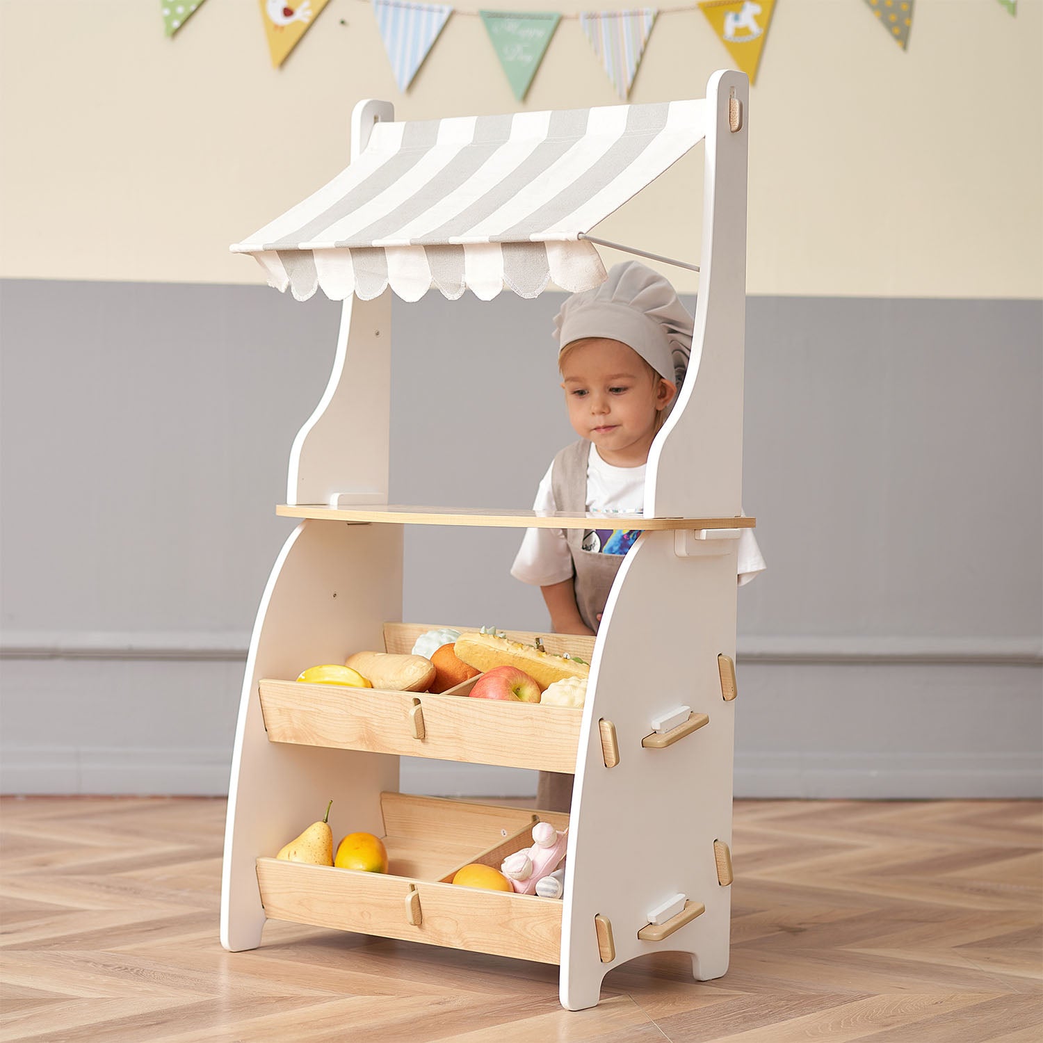 Child playing with a toy fruit stand in a room with a gray wall and wooden play shop.