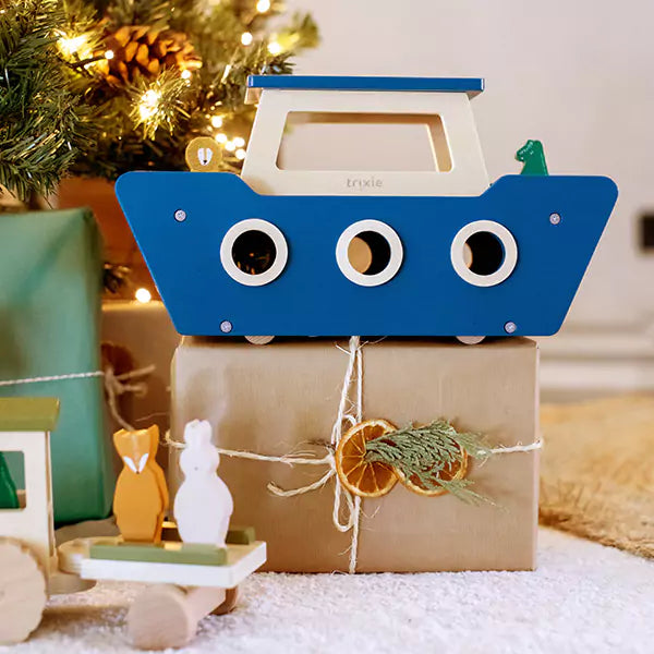  A wooden toy boat sitting on top of a box of presents next to a Christmas tree. This image is from the Kids Stocking Fillers collection.