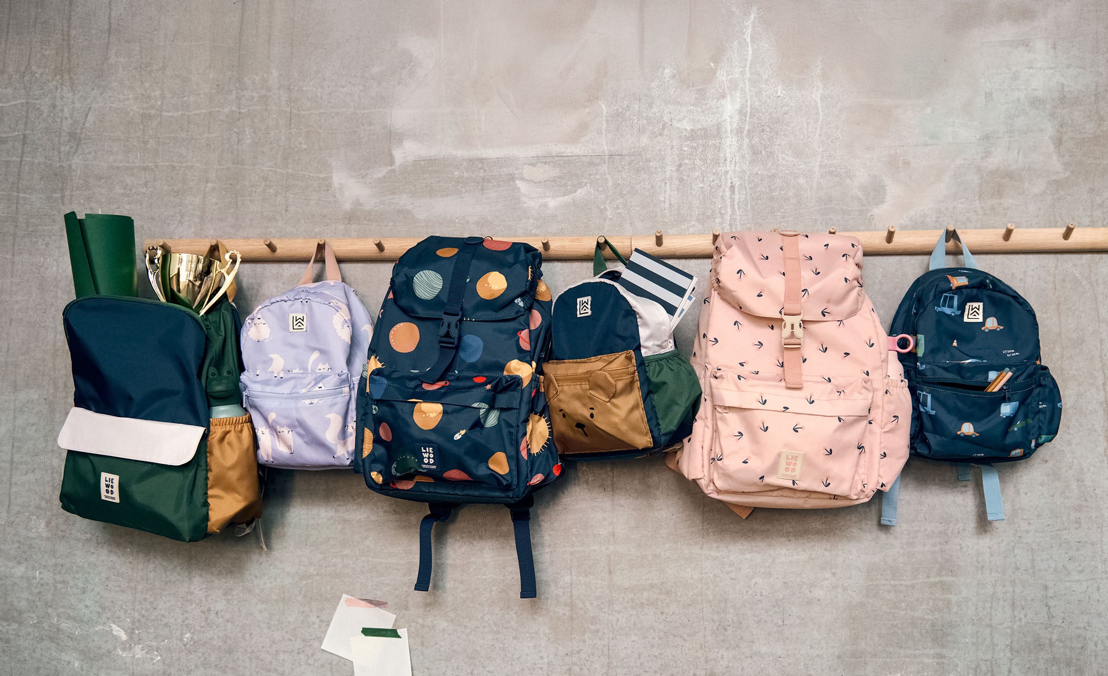 Six colorful backpacks and bags hanging from a wooden rack against a textured gray wall.