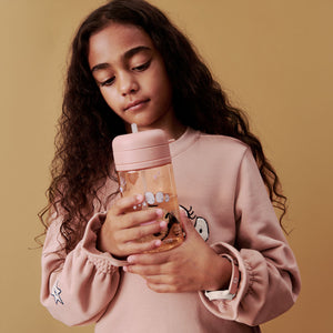 A lifestyle photo of a young girl with long curly hair wearing a pink sweater, holding the peach-colored straw cup close to her chest.