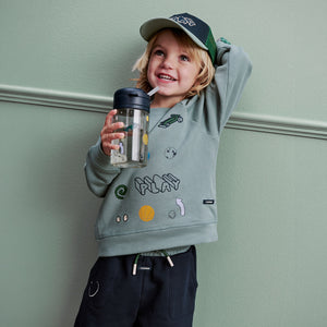A lifestyle photo of a young boy smiling and wearing a green hoodie and cap, holding the play dusty peppermint Liewood straw cup while posing indoors.