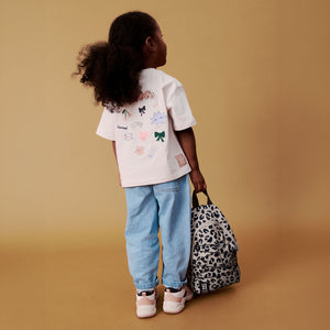 Little girl standing against a warm tan background holding a Liewood leopard print single strap school backpack