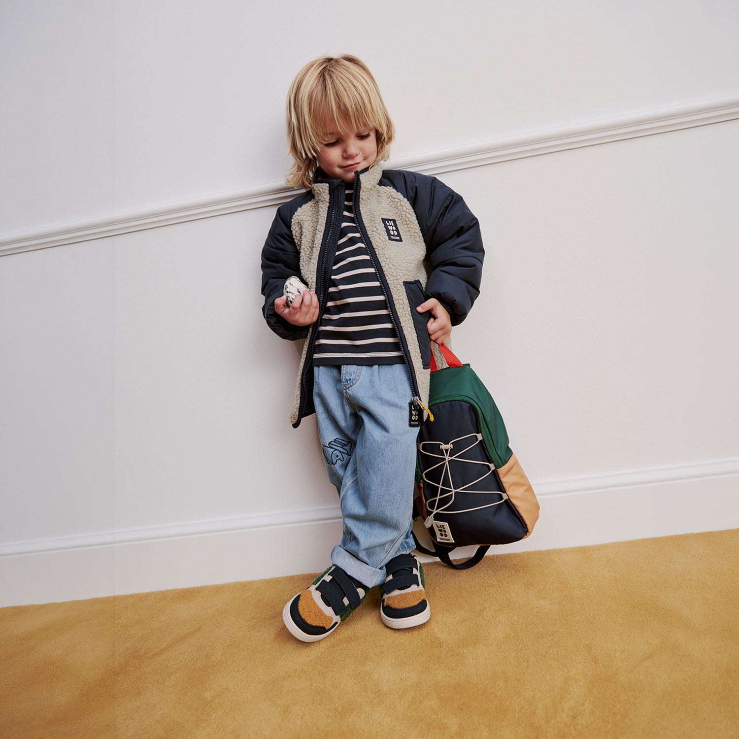 Young boy leaning against a wall wearing a winter jacket and holding a Liewood colorblock single shoulder sling bag