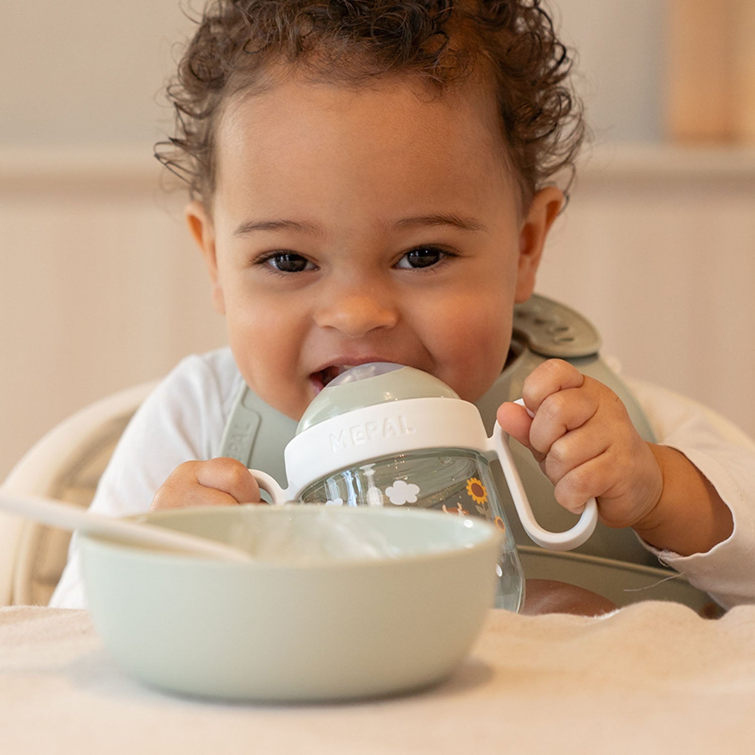A smiling toddler sitting in a highchair independently drinking from the Little Dutch Forest Friends spout cup, comfortably gripping the white side handles.