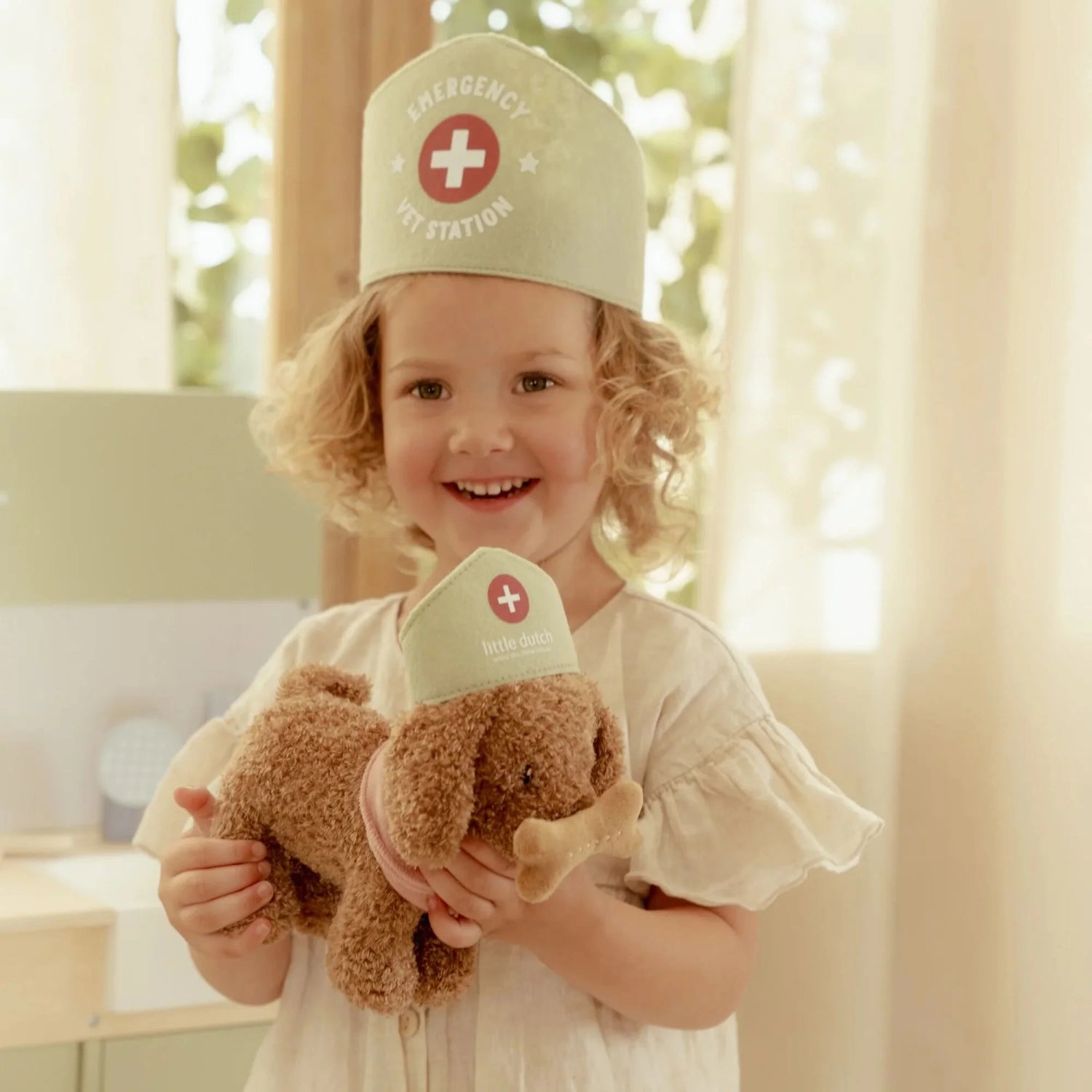 Child wearing a medical-themed cap and holding a plush toy in a bright room.