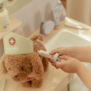 Teddy bear with a 'little dutch' hat being given a tube of toothpaste by a hand in a bathroom setting.