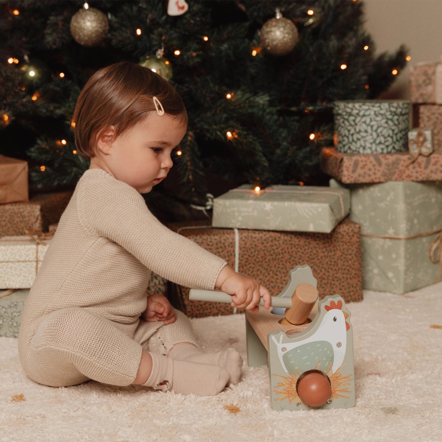 Child playing with a wooden toy in front of a decorated Christmas tree with presents.